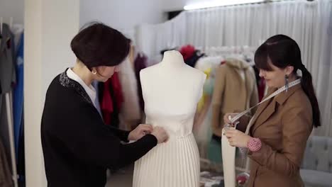 Two-professional-tailors,-designers-working-with-new-model-tailoring-white-dress-on-mannequin-in-studio,-atelier.-Fixing-sleeves-and-pinning-the-skirt.-Fashion-and-tailoring-concept.-Slow-motion