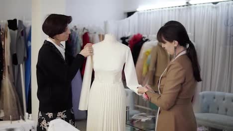 Two-caucasian-female-tailors-collaborating-on-white-wedding-dress-and-pinning-unfinished-garment-on-mannequin-in-fashion-studio,-fixing-sleeves.-Clothes-and-hangers-on-blurred-background