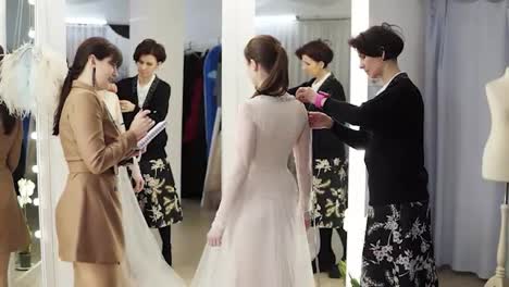 Two-female-dress-designers-and-a-beautiful-bride-trying-her-wedding-gown-in-elegant-boutique-or-tailor's-studio.-A-woman-in-a-white-dress-standing-in-front-the-miror.-Slow-motion