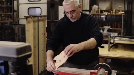 Senior-man-in-black-sweater-grinds-the-fish-shape-pattern-with-grinding-machine-with-sawdust-flying-into-the-sides,-profession,-carpentry-concept.-Caucasian-man-sanding-wood-at-the-workshop.-Slow-motion