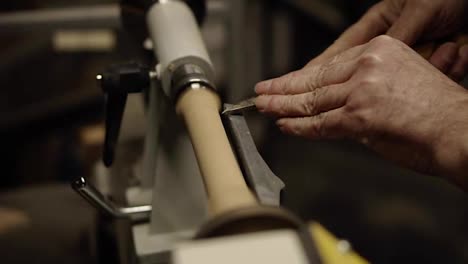 Slow-motion-carpenter-wrinkled-hands-cutting-wooden-knob-out-of-wood-piece-spinning-on-machine-using-chisel,-close-up-shot
