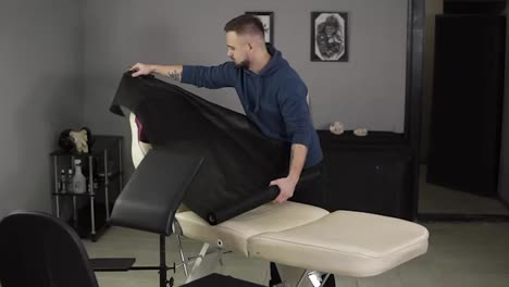 The-procedure-for-preparing-the-cabinet-and-equipment-for-tattooing.-The-caucasian-man-prepares-the-room-and-preparations-to-work.-Covers-the-chair-with-disposable-black-sheet