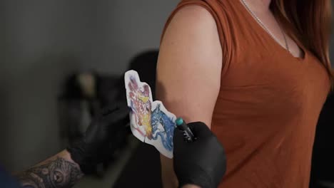 The-tattoo-artist-applies-the-sketch-on-the-woman's-skin-of-upper-hand.-Trying-to-fit-the-colorful-sketch-on-female-unrecognizable-client.-Slow-motion