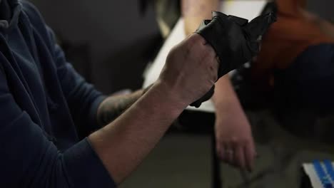 Close-up-of-hands-tattoo-artist-putting-on-the-latex-black-gloves-before-a-tattoo-session-at-the-tattoo-shop.-Tattooist-sitting-on-chair-in-front-the-unrecognizable-client.-Slow-motion