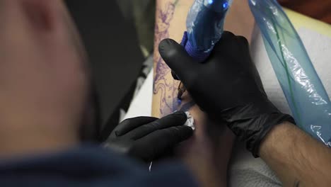 A-close-up-of-a-tattoo-artist-holding-tattooing-gun-and-makes-a-tattoo-on-a-woman's-arm-using-blue-paint.-Professional-tattooist-works-in-studio.-High-angle-footage-from-the-shoulder-of-master