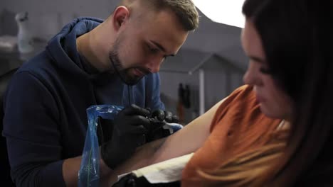 Portrait-of-a-tattoo-artist-demonstrates-the-process-of-getting-colour-tattoo.-Modern-tattoo-parlor.-Bearded-tattoo-artist-and-a-female-client-on-foreground-in-a-beauty-salon