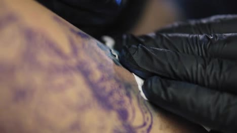 Needle-tattoo-machines-inject-a-blue-ink-into-the-skin-of-a-woman.-Tattoo-art-on-body.-Makes-a-colorful-tattoo.-Professional-tattooist-in-black-gloves-doing-tattooing-in-studio.-Close-up