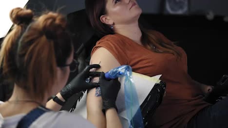 The-tattoo-artist-at-work.-A-red-headed-girl-holding-a-tattoo-machine-and-applied-in-a-pattern-on-the-skin-of-her-female-client.-Blue-ink-in-the-tattoo-machine.-High-angle-footage-from-the-shoulder