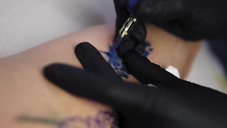 A-close-up-of-a-tattoo-artist-holding-tattooing-gun-and-makes-a-tattoo-on-a-woman's-arm-using-blue-paint.-Professional-tattooist-works-in-studio.-Female-tattoo-master-in-black-gloves