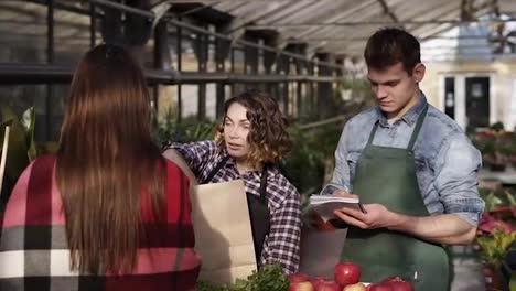 European-saleswoman-wearing-apron-is-giving-organic-food-to-customer-in-greenhouse.-Woman-is-packing-greens,-fruits-and-vegetables-to-a-brown-paper-bag-while-man-making-notes.-People-and-healthy-lifestyle-concept