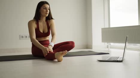 Sportliche-Frau-Macht-Dehnübungen-Auf-Einer-Yogamatte,-Während-Sie-Sich-Online-Fitnessvideos-Auf-Dem-Laptop-Ansieht