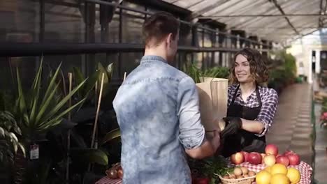 Cheerful-saleswoman-in-black-apron-giving-fresh-vegetables-in-paper-bag-to-customer-in-greenhouse-market.-Smiling-woman-selling-vegetables-to-male-customer.-People-and-healthy-lifestyle-concept