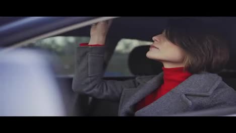 Business-woman-in-grey-coat-and-red-turtleneck-applying-makeup-in-the-car