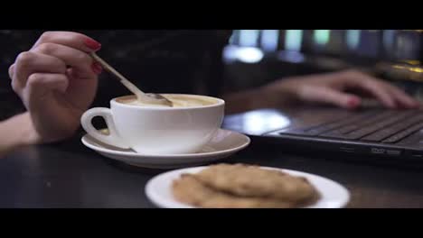 Tasse-Cappuccino-Und-Kekse.-Frau-Mischt-Ihren-Cappuccino-Mit-Einem-Löffel-Und-Arbeitet-Mit-Einem-Laptop-Im-Hintergrund.