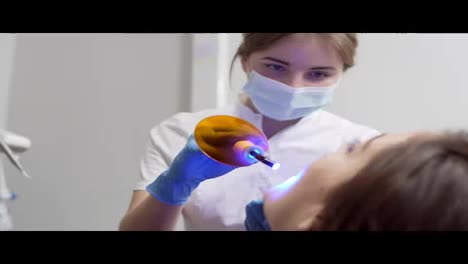 Young-female-dentist-in-mask-and-gloves-using-dental-UV-light-equipment-for-polymer-hardening