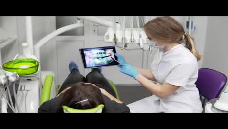 Female-doctor-dentist-is-showing-x-ray-teeth-on-tablet-to-young-female-patient.-Dentist-is-wearing-lab-coat