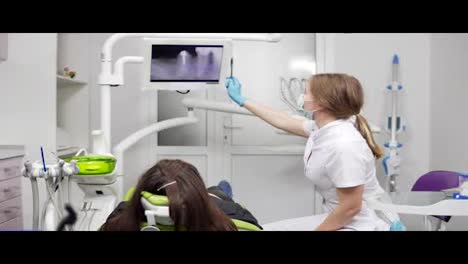 Young-female-dentist-is-showing-x-ray-teeth-on-a-tablet-at-the-chair.-Female-dentist-in-mask-and-lab-coat.-Healthy-teeth-and-dental-healthcare.