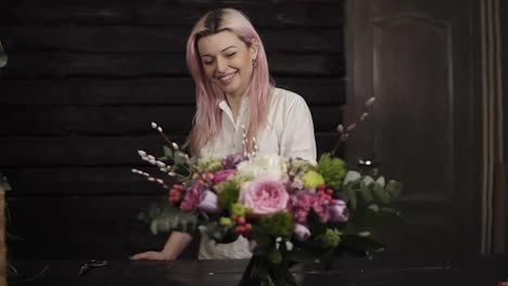 A-beautiful-girl-with-pink-hair-smiles,-admires-a-stunning-bouquet-of-flowers-lying-on-the-surface-opposite-her.-The-girl-is-a-florist.-Dark,-modern-studio