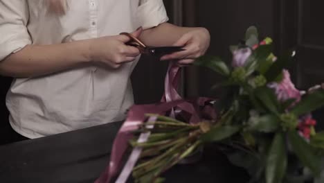 A-girl-in-a-white-shirt-cuts-long-pink-ribbons-and-purple-broad-ribbons-of-guipure-to-decorate-a-bouquet-of-flowers-on-a-table.-Close-up