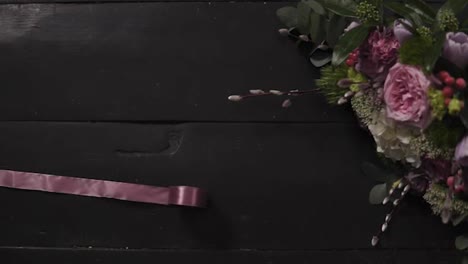 Two-silk-pink-ribbons-sprawled-on-the-dark-wooden-surface,-a-beautiful-bouquet-of-flowers-in-a-frame.-Slow-motion