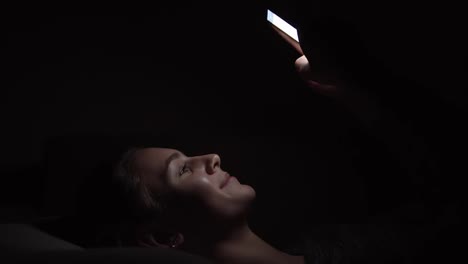 Young-smiling-woman-laying-on-a-couch-and-using-her-phone-in-a-dark-room,-chatting-in-the-darkness-with-smartphone,-communicating-with-friends-online