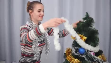Young-happy-woman-in-christmas-sweater-smiling-and-decorating-a-Christmas-tree-hanging-toys.-Shot-in-4k