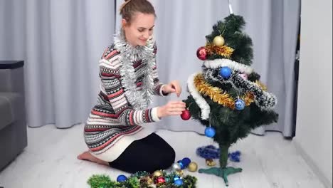 Preparation-for-Christmas-at-home:-Young-happy-woman-decorating-a-Christmas-tree-hanging-toys-and-smiling.-Shot-in-4k