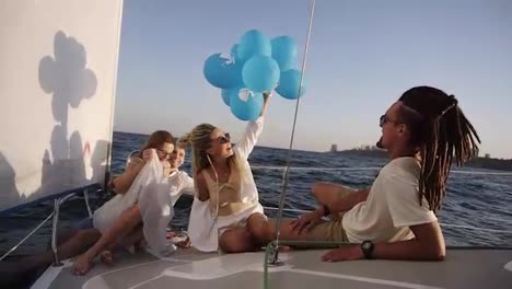 Four-friends-having-relaxing-cruise-on-the-yacht-while-lying-on-a-bow-of-the-boat.-They-enjoying-the-moment---girl-in-a-white-waving-the-balloones,-celebating.-Slow-motion