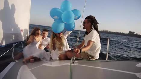 Four-friends-having-relaxing-cruise-on-the-yacht-while-lying-on-a-bow-of-the-boat.-They-enjoying-the-moment---girl-in-a-white-waving-the-balloones,-celebating.-Slow-motion.-Sunny-day-spending-outdoors
