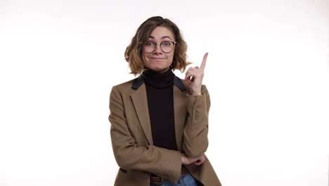 European-pretty-woman-in-stylish-glasses-and-brown-jacket-looks-confused-and-thoughtful-then-gets-an-idea.-Beautiful-young-woman-gets-excited-after-remembering-something,-pointing-up-with-a-finger