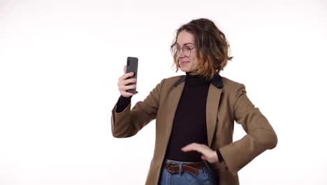 Charming-trendy-young-woman-with-curly-short-hair-having-video-chat-through-phone-and-laughing,-talking-standing-isolated-on-white-background.-Wearing-brown-jacket-and-eyeglasses