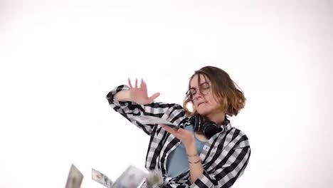 Slow-motion-of-beautiful-curly-short-haired-girl-winning-a-lot-of-money-and-throwing-dollar-bills-in-the-air-from-hands-isolated-over-white-background-in-studio.-Wearing-plaid-shirt,-eyeglasses-and-headphones-on-neck