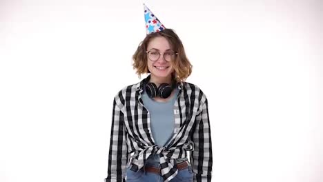 Portrait-of-excited-young-woman-in-plaid-shirt-and-headphones-on-neck-in-birthday-colorful-hat-blow-in-pipe,-falling-confetti-isolated-over-white-background-in-studio.-People-sincere-emotions,-holiday-lifestyle-concept.-Celebrating-party-day