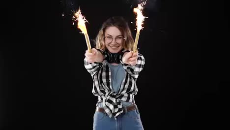 European-hipster-girl-in-black-and-white-plaid-shirt,-headphones-on-neck-celebrating-with-sparklers-and-bengal-fire.-Happy-young-woman-enjoying-and-having-fun-against-black-background.-Slow-motion
