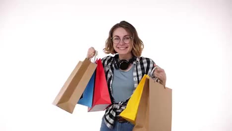 Cheerful-beautiful-european-young-woman-in-glasses-and-headphones-on-neck-holding-colorful-shopping-bags-in-season-sale,-happily-show-them-on-outstretched-hands-isolated-on-white-background