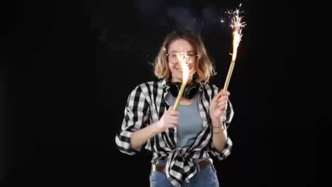 European-hipster-girl-in-black-and-white-plaid-shirt,-headphones-on-neck-celebrating-with-sparklers-and-bengal-fire.-Happy-young-woman-having-fun-and-playfully-dancing-against-black-background.-Slow-motion