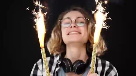 Close-up-european-hipster-girl-in-black-and-white-plaid-shirt,-headphones-on-neck-celebrating-with-sparklers-and-bengal-fire.-Happy-woman-smiling-girl-holds-two-sparklers-close-to-the-camera-against-black-background.-Slow-motion