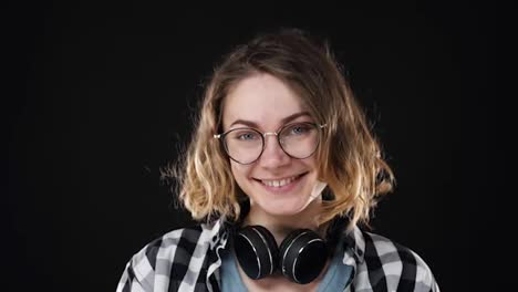 Close-up-portrait-of-young-european-girl-smiling-humbly-into-camera-over-black-background.-Cheerfully-smiling.-Wearing-eyeglasses-and-headphones-on-neck