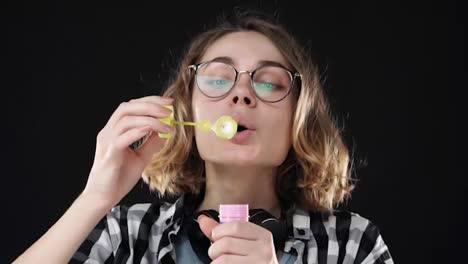 Closeup-portrait-of-happy-emotional-girl-blowing-a-big-soap-bubble-with-stick-on-black-background.-Studio-footage-of-brunette-with-short-hair-and-sensual-lips-wearing-headphones-on-neck