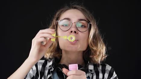 Closeup-portrait-of-happy-emotional-girl-blowing-a-lot-of-soap-bubbles-with-stick-on-black-background.-Studio-footage-of-brunette-with-short-hair-and-sensual-lips-wearing-headphones-on-neck.-SLOW-MOTION