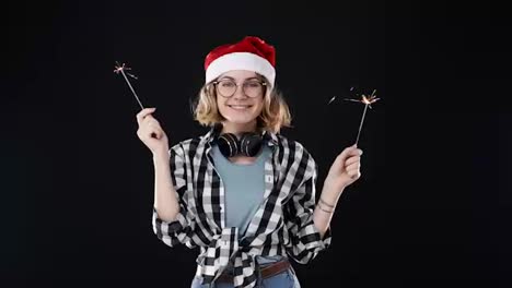 European-hipster-girl-in-black-and-white-plaid-shirt,-headphones-on-neck-and-red-santa-hat-celebrating,-holding-two-bengal-fires.-Happy-woman-smiling-and-gesturing,-looking-to-the-camera-against-black-background.-Slow-motion
