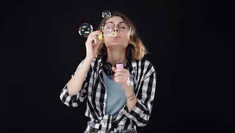 Portrait-of-happy-girl-in-glasses-blowing-a-lot-of-soap-bubbles-with-stick-on-black-background.-Studio-footage-of-brunette-with-short-hair-and-sensual-lips-wearing-headphones-on-neck.-SLOW-MOTION