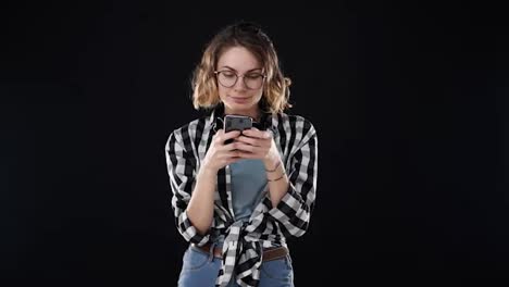 Portrait-of-concentrated-european-woman-wearing-casual-clothing---plaid-shirt-and-jeans,-holding-smartphone-and-typing-text-message-or-chatting,-isolated-over-black-background.-Concept-of-emotions,-friendship,-technology
