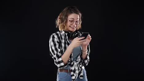 Cheerful-woman-wearing-casual-clothing---plaid-shirt-and-jeans,-holding-smartphone-and-scrolling-the-social-meadia-or-chatting,-isolated-over-black-background.-Getting-excited-with-news-she-reads