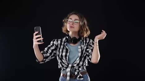 Cute-young-woman-in-plaid-shirt-and-headphones-on-neck-prapering-herself,-looking-to-her-smartphone-and-taking-a-selfie-isolated-on-black-background-in-studio.-Grimacing-face,-showing-tongue