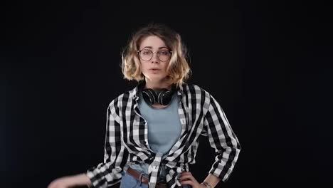 Serious-young-woman-in-black-and-white-plaid-shirt-and-headphones-on-neck,-doing-NO-gesture,-disappointed-gesturing-isolated-on-black-background-in-studio.-People-sincere-emotions
