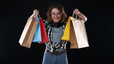 Cheerful-beautiful-european-young-woman-in-glasses-and-headphones-on-neck-holding-colorful-shopping-bags-in-season-sale,-happily-show-them-on-outstretched-hands-isolated-on-black-background