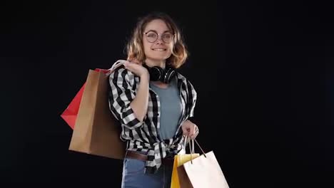 Smiling-excited-buyer-young-modern-woman-with-headphones-on-neck-and-stylish-glasses-posing-with-colorful-paper-shopping-bags,-holding-on-shoulder-isolated-at-black-studio-background