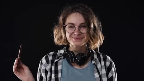 Close-up-brunette-young-woman-with-glasses-and-headphones-posing-playfully-eat-hold-in-hand-chocolate-bar-with-nuts-isolated-over-black-background-in-studio.-People-sincere-emotions-lifestyle-concept