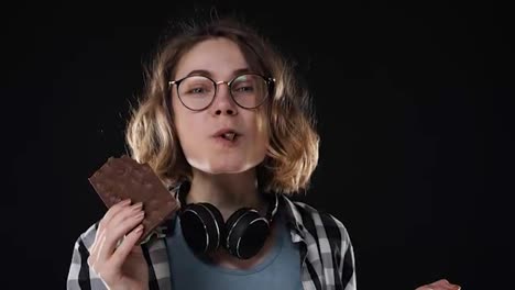 Close-up-brunette-young-woman-with-glasses-and-headphones-posing-playfully-eat,-chewing-chocolate-bar-with-nuts-isolated-over-black-background-in-studio.-People-sincere-emotions-lifestyle-concept